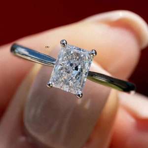 May include: A silver ring with a large, clear, rectangular-shaped diamond. The diamond is set in a prong setting. The ring is on a finger.