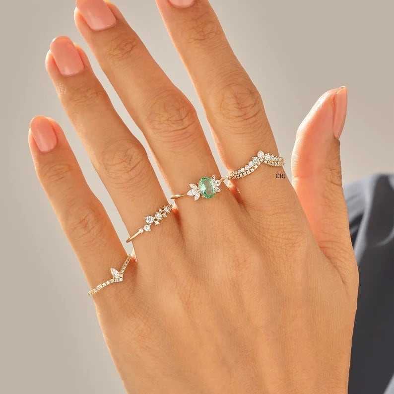 May include: A set of four gold rings with small clear stones and a green oval stone. The rings are worn on a hand with pink nail polish.