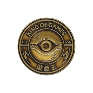 May include: A gold-colored metal pin with the words "King of Games" and "Est. 1996" around a stylized eye with lightning bolts. The text "遊戯王" is written in Japanese below the eye.
