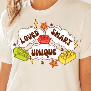 May include: White t-shirt with a graphic design featuring the words "Loved", "Smart", and "Unique" in a retro font. The design also includes colorful cartoon building blocks and stars.