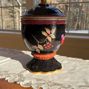 May include: A black ceramic jar with a lid, featuring hand-painted floral designs in red, pink, and green. The jar has an orange base and a decorative finial on the lid. The jar is sitting on a white lace tablecloth.