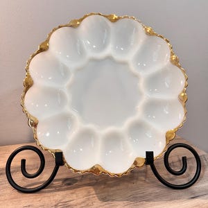 May include: White egg plate with a gold rim and 12 individual egg-shaped depressions. The plate is on a black metal stand.