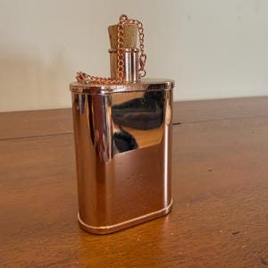 Op de afbeelding: Een gepolijste koperkleurige metalen heupfles met een ovale basis en een reflecterend oppervlak. De fles heeft een cilindrische hals met een lichtbruine kurkstop. Een dunne koperkleurige metalen ketting is aan de hals van de fles bevestigd.