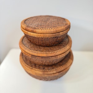 May include: Three round, woven baskets stacked on top of each other. The baskets are made of a light brown material and have lids. The baskets are of varying sizes, with the largest at the bottom and the smallest at the top.