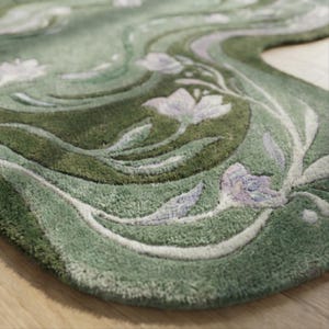 Op de afbeelding: Een groene vloerkleed met een bloemenontwerp. Het vloerkleed heeft een wervelend patroon in groentinten, met witte en lavendelkleurige bloemen en bladeren. Het vloerkleed heeft een zacht, getextureerd uiterlijk.