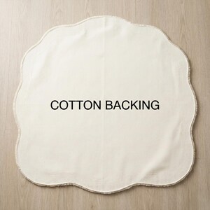 May include: Off-white, scalloped-edge cotton mat with the text "COTTON BACKING" in black. The mat is on a light wood surface. The mat's shape is reminiscent of a cloud or flower.
