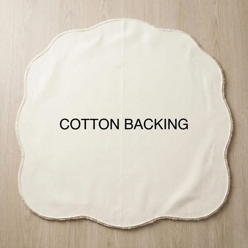May include: Off-white, scalloped-edge cotton mat with the text "COTTON BACKING" in black. The mat is on a light wood surface. The mat's shape is reminiscent of a cloud or flower.