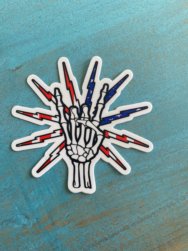 Op de afbeelding: Een sticker met een skelet hand die het 'rock on' gebaar maakt. De hand is omringd door bliksemschichten, en het ontwerp is in de kleuren van de Amerikaanse vlag.