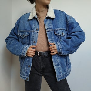 Op de afbeelding: Een blauw spijkerjack met een witte shearling kraag. De jas heeft twee borstzakken en een rood Levi's label. De persoon draagt een bruine coltrui, een zwarte jeans en een zwarte riem.