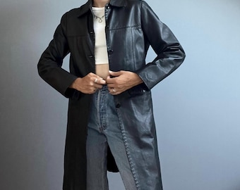 May include: A black leather trench coat featuring a classic collar, long sleeves, and a button-down front closure. The coat has a smooth texture and a structured fit, extending to mid-thigh length, worn over a white crop top and blue denim jeans.