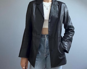 Könnte beinhalten: Ein schwarzer Lederblazer mit fallendem Revers, langen Ärmeln und einer Knopfleiste mit drei Knöpfen. Die Jacke wird über einem weißen bauchfreien Oberteil und einer hellblauen Jeans getragen und zeigt eine strukturierte Passform.