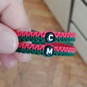 Könnte beinhalten: Zwei handgefertigte Makramee-Armbänder. Jedes Armband ist rot und grün mit einer schwarzen Perle in der Mitte. Eine Perle hat den Buchstaben "C" und die andere den Buchstaben "M". Die Armbänder sind wahrscheinlich für Paare oder Freunde.