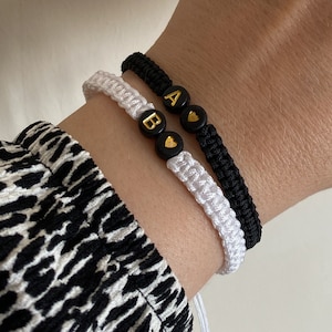 Op de afbeelding: Drie macramé-armbanden, twee zwarte en één witte, worden getoond. De zwarte armband heeft gouden letterkralen met de letters "A" en een hart. De witte armband heeft een gouden letterkraal met de letter "B".