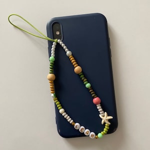 Può includere: Un laccio per telefono verde con perline colorate, tra cui perline bianche, verdi, marroni e rosa. Il laccio ha il nome "LEAH" scritto in perline bianche con lettere dorate. Un ciondolo a forma di stella marina bianca è attaccato al laccio.