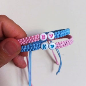 Op de afbeelding: Twee vriendschapsarmbanden met roze en blauwe draad, elk met een witte kraal met een letter en een hartbedel. De armband aan de linkerkant heeft een witte kraal met de letter "B" en een roze hartbedel. De armband aan de rechterkant heeft een witte kraal met de letter "K" en een blauwe hartbedel.