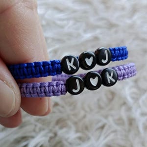 May include: Two handmade friendship bracelets. One is blue, the other is purple. Each bracelet has black beads with white letters and a heart symbol. The blue bracelet has the letters "K", a heart, and "J". The purple bracelet has the letters "J", a heart, and "K".