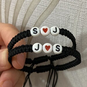 Op de afbeelding: Twee zwarte vriendschapsarmbanden met witte kralen die "S♥J" en "J♥S" spellen.