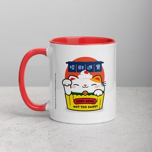 May include: White ceramic mug with a red rim and handle. The mug features a cartoon illustration of a white cat with orange patches, holding a yellow box with the text "Very Good Not Too Sweet". The cat is wearing a green collar and has a cherry on its head. The background features a red circle with three blue and white banners with Japanese characters.