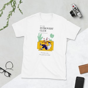 May include: White t-shirt with a graphic design of a person sitting in a box with plants. The text on the shirt reads "EST. 2022 INTROVERT CLUB Escape & Unwind - Members only - No guests permitted"