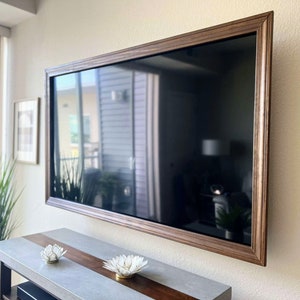Puede incluir: Un televisor rectangular grande con una pantalla oscura está montado en una pared beige. El televisor tiene un marco de madera marrón ancho y ornamentado. Debajo, una consola gris y marrón con esculturas de flores blancas decorativas.