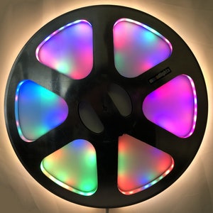 Op de afbeelding: Een zwarte haspelbandspeler met een cirkelvormig ontwerp en vijf verlichte secties. Elke sectie heeft een andere kleur, waaronder roze, paars, blauw en groen. De tekst "moodlights" is op de speler gedrukt.