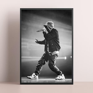 May include: Black and white framed print of a person performing on stage, holding a microphone. The person is wearing a hooded jacket, jeans, and sneakers. The background is blurred with stage lights.