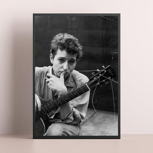 Könnte beinhalten: Schwarz-Weiß-Druck in einem Rahmen, der einen Musiker mit Gitarre und Stift zeigt. Der Musiker, der ein Jeanshemd trägt, blickt in die Kamera. Die Gitarre trägt den Markennamen "Gibson" auf der Kopfplatte.