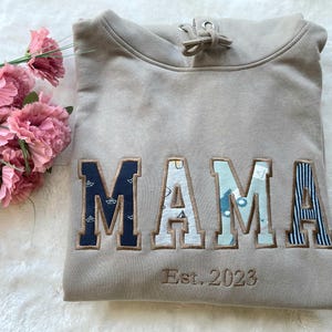 Könnte beinhalten: Ein hellbraunes Kapuzenpullover mit dem Wort "MAMA" in großen, bunten Buchstaben. Die Buchstaben bestehen aus verschiedenen gemusterten Stoffen. Der Text "Est. 2023" ist unter dem Wort "MAMA" gestickt.