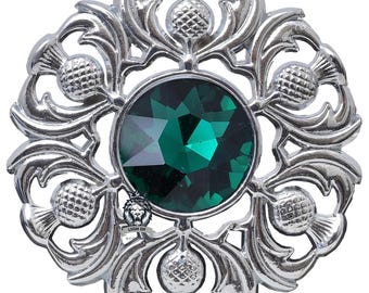 May include: A silver-toned brooch with a central, faceted, emerald-green gemstone. The brooch features a detailed, openwork design of thistles and leaves, with a circular arrangement. The Lion of Scotland logo is visible at the bottom.