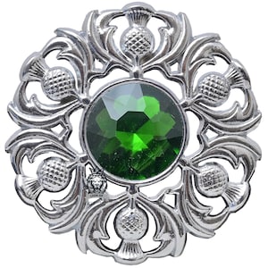 Op de afbeelding: Zilverkleurige broche met een centrale, ronde, gefacetteerde groene edelsteen. De broche heeft een gedetailleerd, opengewerkt ontwerp van gestileerde distels en bladeren, waardoor een cirkelvormig patroon ontstaat. Het ontwerp is symmetrisch en sierlijk.