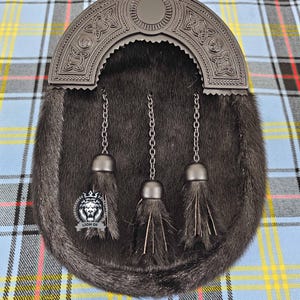 May include: A dark gray sporran with a decorative metal cantle featuring Celtic knotwork. The sporran has a fur body, three hanging chains with tassels, and a logo. The background is a blue, black, red, and yellow plaid.