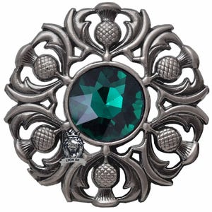 May include: A silver-coloured brooch with a central, faceted emerald-green stone. The brooch features a detailed design of thistles and leaves, with a small emblem at the bottom. The design is circular and ornate.
