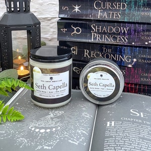 Puede incluir: Dos velas con la etiqueta "Seth Capella" y el texto "This candle smells like Earthy Woods | Musk | Moon Stone" sobre una pila de libros con títulos como "Cursed Fates", "Shadow Princess" y "The Reckoning".