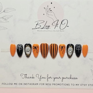 May include: A set of ten almond-shaped press-on gel nails featuring a Halloween theme with orange and black colors. Designs include vertical stripes, spider webs, bows, and ghost illustrations on a display card with the text Bling It On.