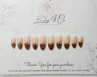 May include: A set of ten almond-shaped press-on gel nails featuring a nude base with hand-painted brown pumpkin designs at the tips. The nails are displayed on a white card with the text Bling It On Handcrafted Press On Gel Nails.