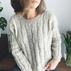 Könnte beinhalten: Ein hellbeige Zopfmuster-Pullover mit Rundhalsausschnitt. Der Pullover hat vertikale Zopfmuster und lange Ärmel. Die Person trägt dunkelgraue Jeans. Der Hintergrund ist eine neutrale weiße Wand und ein Holztisch mit einer Blumenvase.