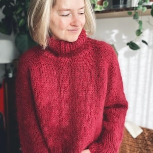 Może przedstawiać: Zbliżenie na kobietę w czerwonym swetrze z golfem. Sweter jest dziergany luźnym, fakturowanym ściegiem. Kobieta ma na sobie szare dżinsy i blond włosy.