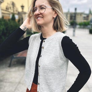 May include: A light grey knitted waistcoat with a button fastening. The waistcoat is cropped and has a relaxed fit. The model is wearing the waistcoat over a black long-sleeved shirt and brown trousers.