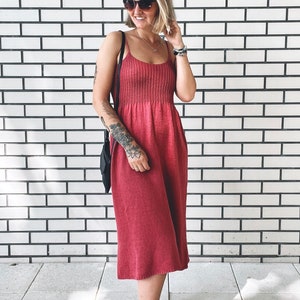 May include: A woman wearing a red midi dress with a thin strap and a gathered bodice. She is wearing sunglasses and a necklace. The dress has a relaxed fit and is made of a soft, comfortable fabric.