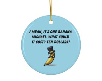 May include: A circular, pale blue ceramic Christmas ornament with a gold hanging cord. It features a cartoon banana wearing a top hat and mustache. The text on the ornament reads, "I MEAN, IT'S ONE BANANA, MICHAEL. WHAT COULD IT COST? TEN DOLLARS?"