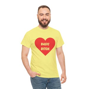 May include: Yellow t-shirt featuring a red heart with the text "BASIC BITCH." The casual top is a simple design, suitable for everyday wear. The text is in a contrasting font.