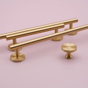 May include: Four gold-toned cabinet pulls with a circular, textured design. The pulls are arranged in a staggered pattern on a pink background.
