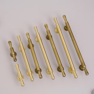 May include: A collection of brass-toned door pulls in various sizes. The pulls have a cylindrical design with rounded ends and circular mounting plates. The finish varies from bright gold to a darker, aged brass. The hardware is arranged on a light gray surface.