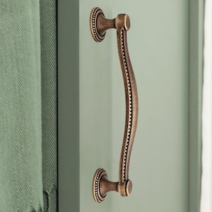 May include: A curved, ornate cabinet door pull with a beaded design. The pull is made of metal and has a bronze finish.