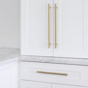 May include: White kitchen cabinets with gold-coloured hardware. The upper cabinets have long, vertical pulls, while the lower cabinets and drawers feature horizontal pulls. The countertop is white with grey veining.