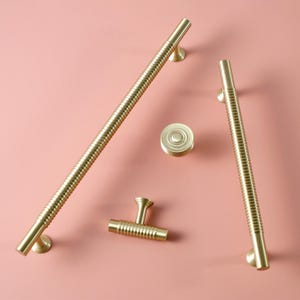 May include: A set of gold-toned cabinet hardware, comprising two long bar pulls, a small knob, and a T-shaped pull. The hardware features a ribbed design and is displayed on a pink backdrop. The bar pulls are approximately 30 cm in length.