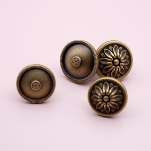 Puede incluir: Cuatro tiradores de armario de bronce antiguo. Dos tiradores tienen un dise&ntilde;o floral y dos tienen un dise&ntilde;o circular con una peque&ntilde;a flor en el centro. Los tiradores est&aacute;n sobre un fondo rosa claro.