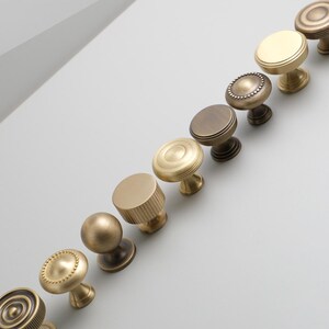 May include: A collection of 10 brass cabinet knobs in various styles and finishes, including round, square, and cylindrical shapes. The knobs are arranged in a row on a white surface.