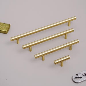 Può includere: Quattro maniglie per mobili color ottone di diverse lunghezze. Le maniglie sono cilindriche con una base a forma di T. La maniglia più lunga misura circa 30 cm, e la più corta circa 5 cm. Le maniglie sono disposte diagonalmente.