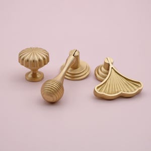 May include: Three gold-toned cabinet knobs with different designs. One is a round knob with a fluted design, another is a round knob with a fluted design and a long, fluted handle, and the third is a leaf-shaped knob with a fluted design.